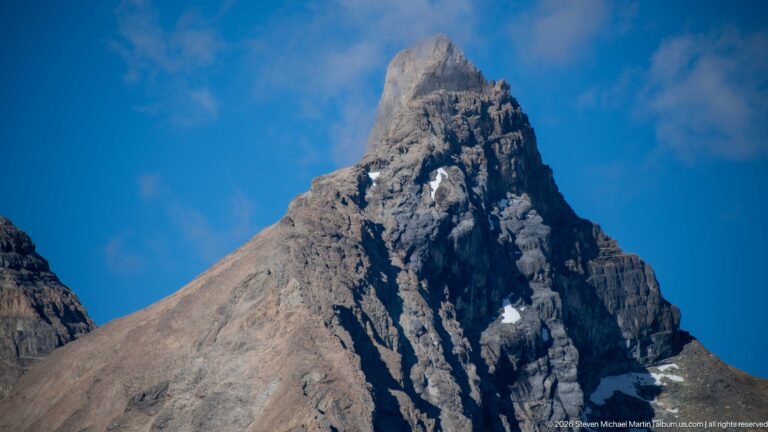 A dark rocky spire rises above a pale scree shoulder against clear blue sky.
