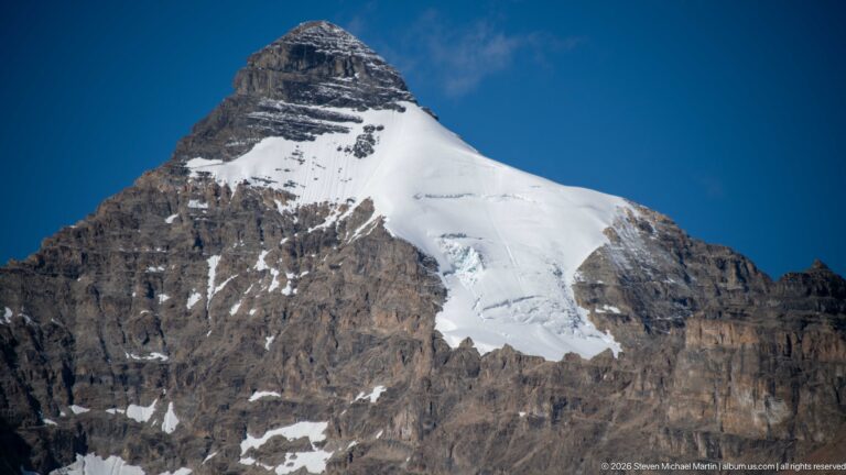 A broad snowfield and fractured icefall cover a steep, horizontally banded mountain face.