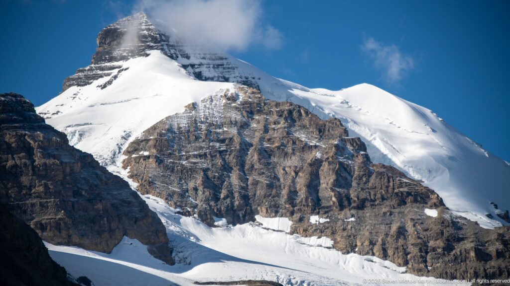 A bright snowfield and broken glacier cover a steep mountain above exposed rock bands.