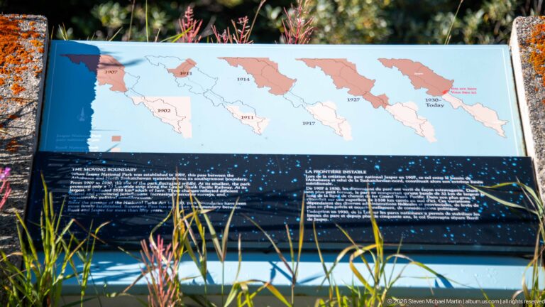 A blue interpretive panel displays dated glacier-boundary maps above explanatory text and foreground grasses.