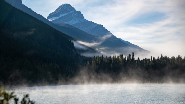 Blue lake water lies below a dark forested mountain as cloud and mist drift across its slope.