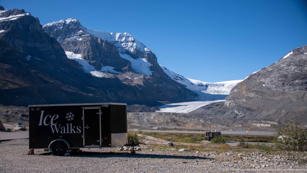 A black Ice Walks trailer is parked on gravel below a glacier between steep mountain walls.