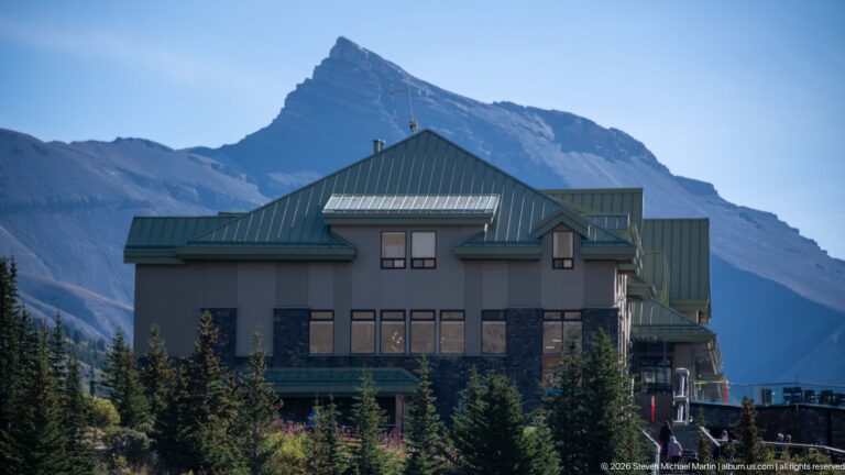 A green-roofed lodge stands among conifers beneath a steep layered mountain peak.