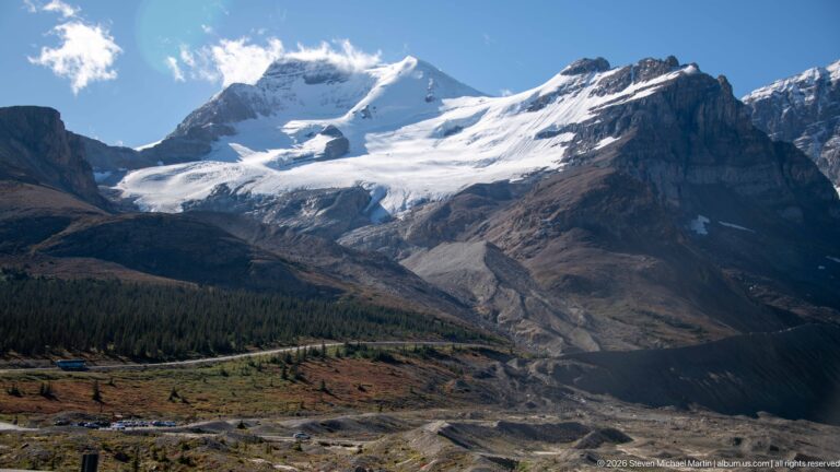 A broad glacier flows into a debris-covered valley bordered by forest and steep mountains.