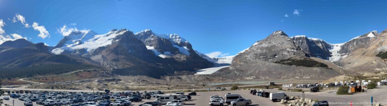 A broad glacier descends between rocky peaks above a parking area filled with cars, buses, and recreational vehicles.