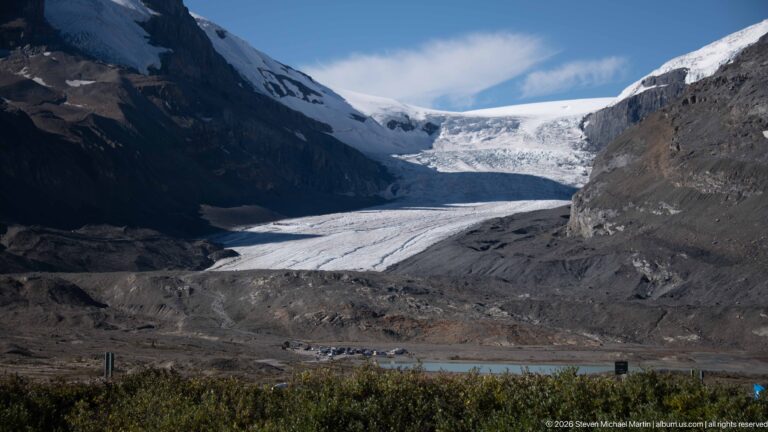 A broad glacier descends between dark rock walls to a gravelly terminus and moraine.