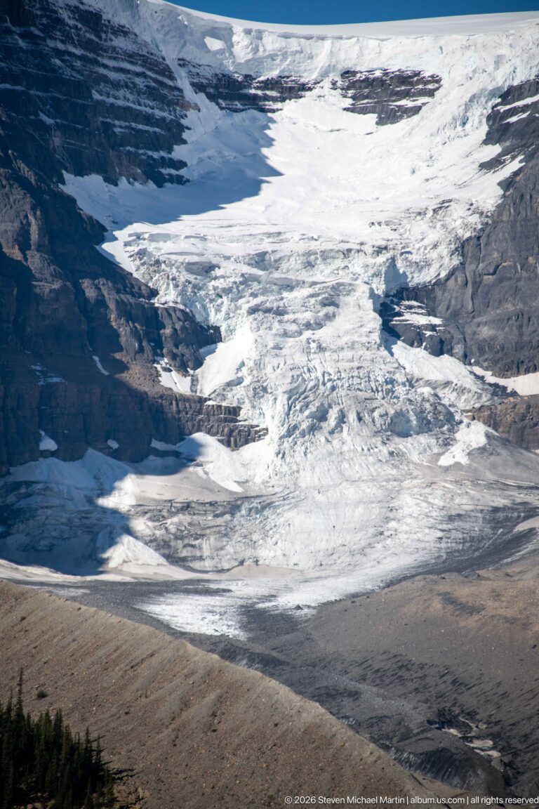 Bright ridges and dark crevasses pattern the rough surface of a glacier beneath a mountain slope.