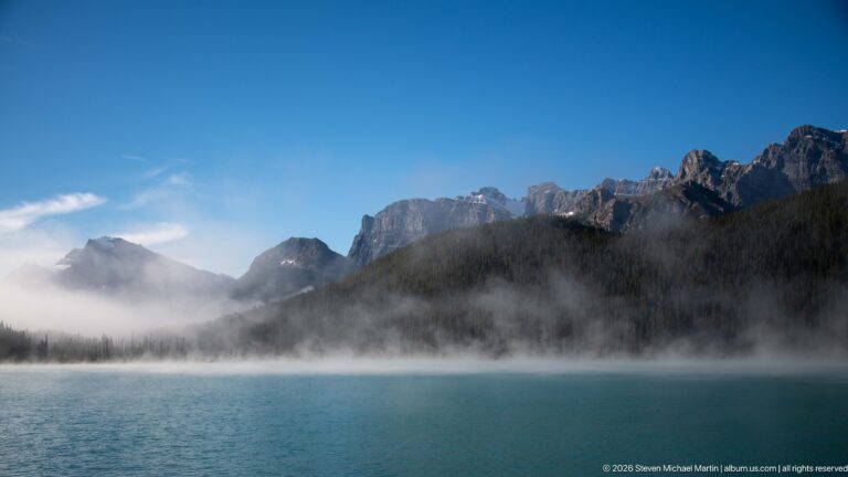 Dark peaks rise above a low fog bank and forested shore across a deep blue lake.