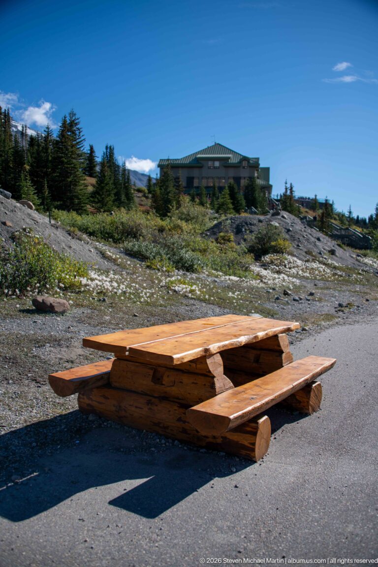 A timber picnic table sits on pavement below a mountain visitor building and alpine vegetation.