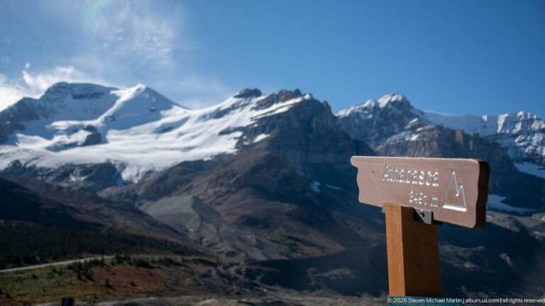 A Mount Athabasca sign stands before a glacier flowing between steep peaks toward a rocky terminus.