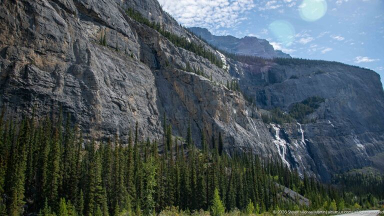 Thin waterfalls cross a receding cliff above evergreen forest under bright blue sky.