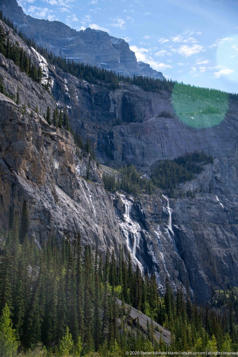 Several bright streams cross a broad stepped cliff above a dark conifer forest.