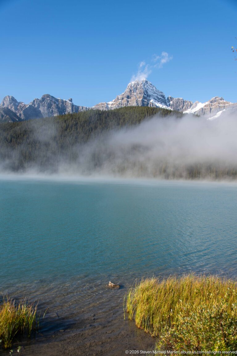 A broad blue lake lies below a pale ridge softened by fog, with grasses along the near shore.