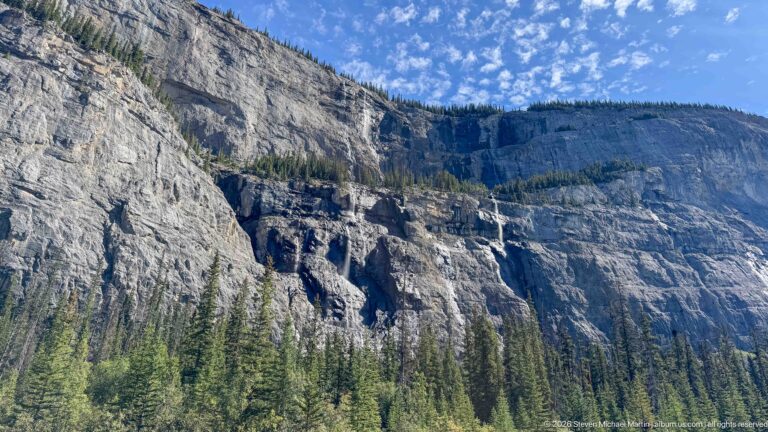 A long limestone escarpment with scattered waterfalls rises above a dense conifer slope.