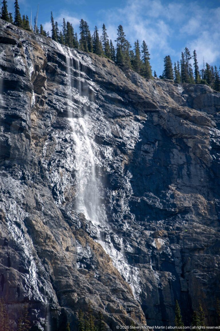 Meltwater splashes over a sloping dark rock shelf beneath a line of trees.