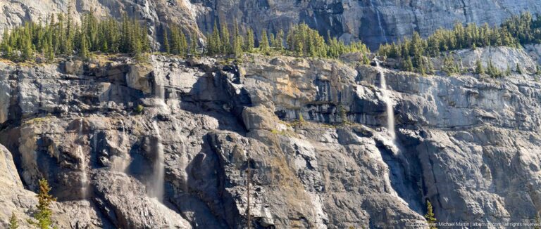 Thin waterfalls descend separate ledges on a tall pale limestone cliff topped by spruce.