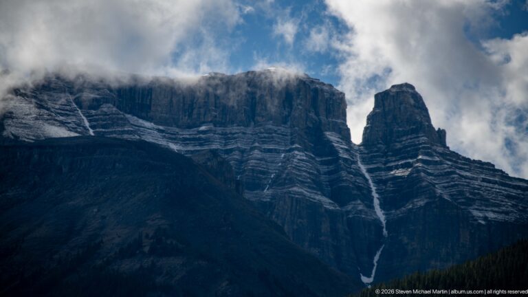 A narrow waterfall descends a tall layered cliff beneath drifting cloud, above a dark forested slope.