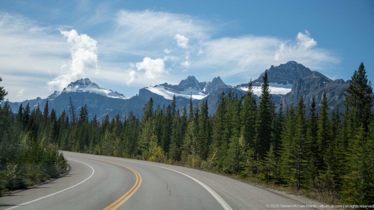 A two-lane mountain highway curves through conifers toward jagged, snow-dusted peaks under blue sky.
