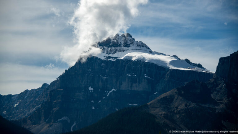 Cloud partly veils a snowy summit above dark cliffs and a glacier-filled saddle.
