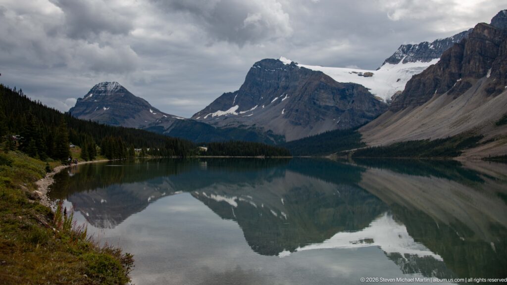 A pale glacier lies in a notch between dark peaks above Bow Lake and its forested shoreline.