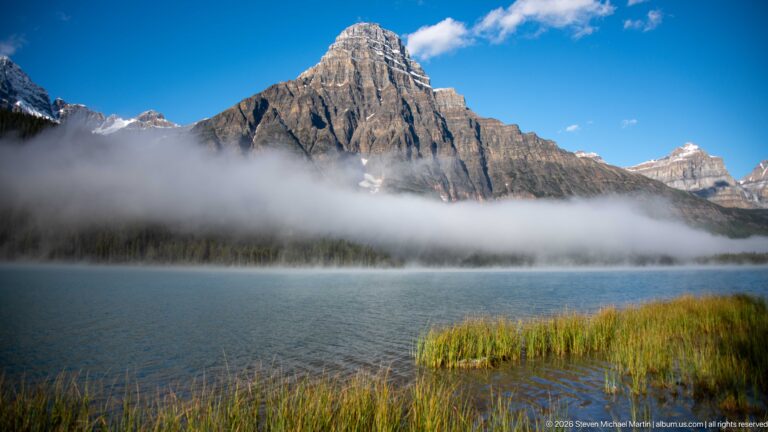 A sharp mountain peak rises above fog and forest across a blue lake edged with reeds.