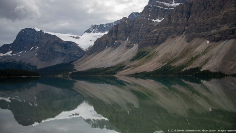 A glacier descends between layered rock walls, reflected in a dark mountain lake.