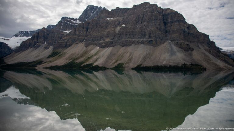A steep layered mountain and pale scree fan are mirrored in Bow Lake’s nearly still surface.