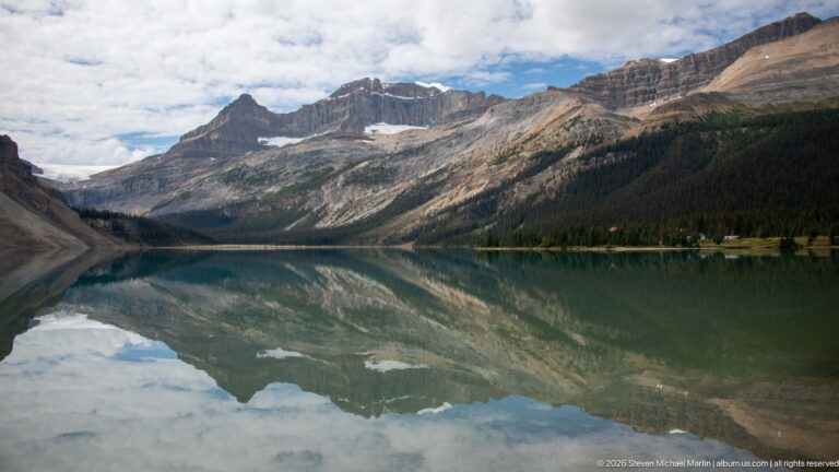 A broad lake extends toward a mountain gap, reflecting forested slopes and clouds beneath a blue sky.
