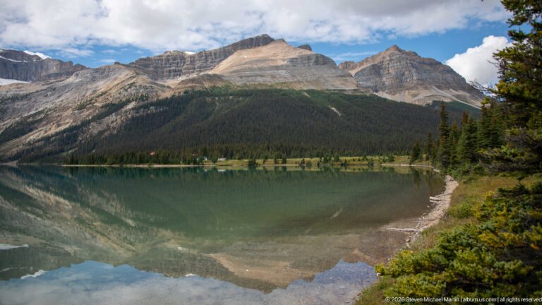 A curved gravel shore borders a reflective alpine lake beneath layered, partly snow-covered peaks.