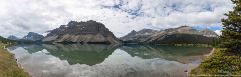 Jagged mountains and patchy snow rise above Bow Lake, with their cloud-dappled slopes reflected in still water.