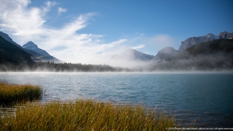 Low mist stretches across a blue lake between dark forested slopes and grasses at the near shore.