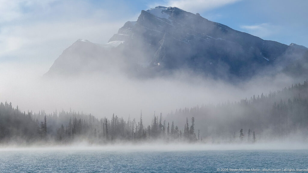 Blue lake water fills the foreground beneath a fog bank and dark mountain ridge.