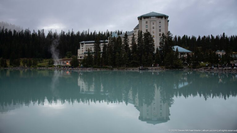 The Fairmont Chateau Lake Louise tower and surrounding trees reflect in still turquoise lake water.