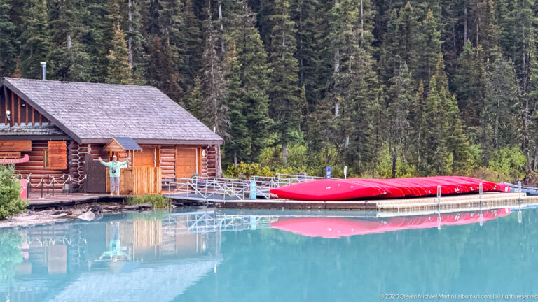 A visitor stands by a log boathouse and red canoes reflected in Lake Louise.
