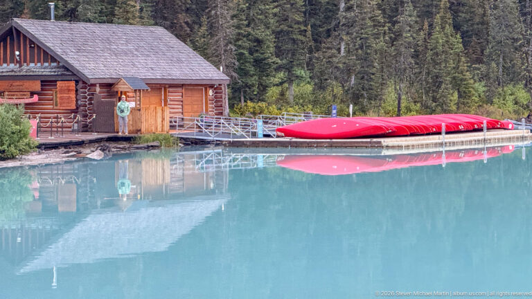 Red canoes rest on a dock beside a log boathouse, reflected in turquoise water beneath evergreen forest.