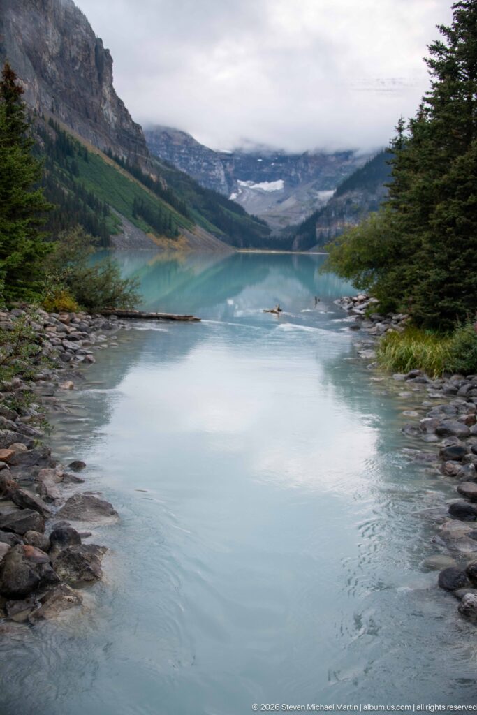 Pale turquoise water flows past rocky banks and a narrow strip of land beneath an overcast sky.