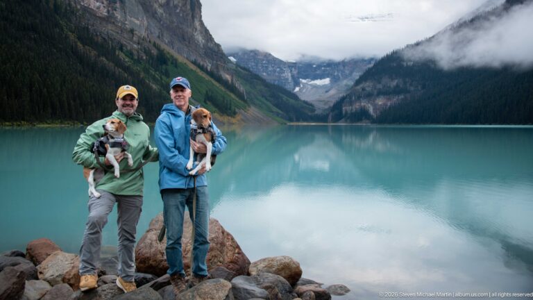 Two visitors with small dogs stand at the left of a wide view across Lake Louise.