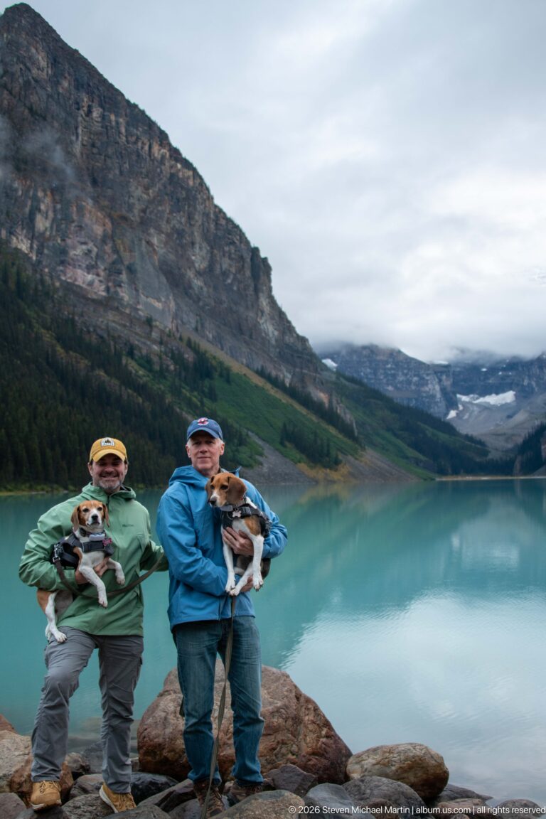 Two visitors hold small dogs among shoreline boulders, with turquoise Lake Louise and its mountain valley behind them.