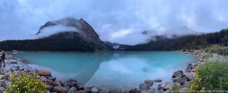 Rounded rocks border Lake Louise beneath a forested mountain and cloud-covered ridges.