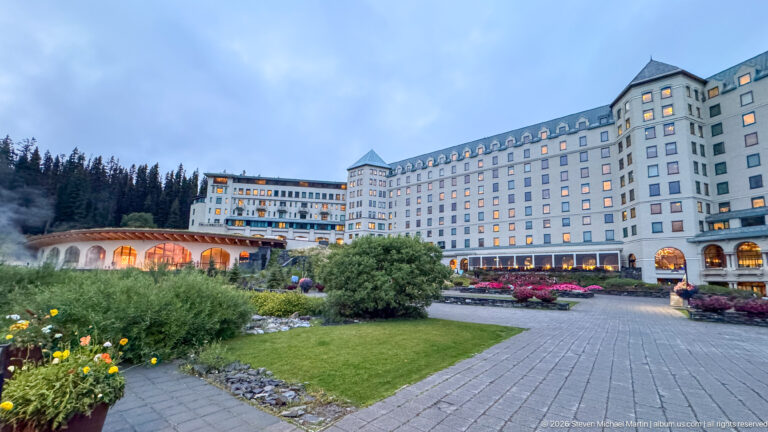 The full Fairmont Chateau Lake Louise stretches beyond a lawn and curved garden path beneath a pale evening sky.