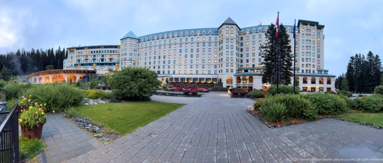 The Fairmont Chateau Lake Louise rises above a formal lawn, curved path, and colorful flower beds at dusk.