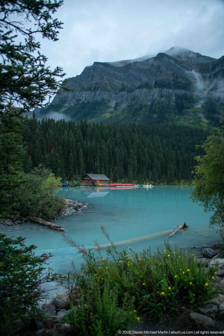 Red canoes line a far-shore dock at Lake Louise, reflected in calm water below a rocky, forested slope.