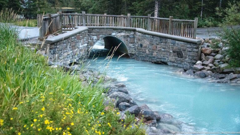A milky turquoise stream passes through a rounded opening beneath a stone footbridge with timber railings.