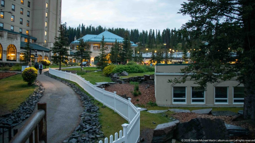 A white fence curves through the landscaped grounds toward the warmly lit Fairmont Chateau Lake Louise at dusk.