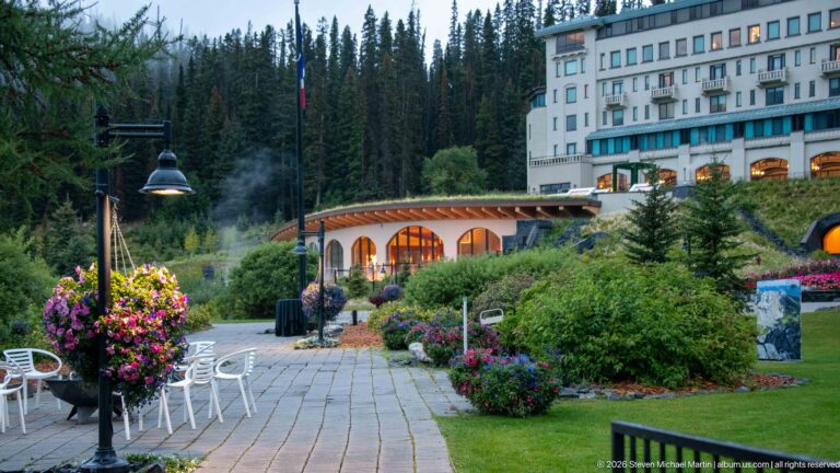 A paved garden terrace with flower baskets leads toward the illuminated arcade of the Fairmont Chateau Lake Louise.