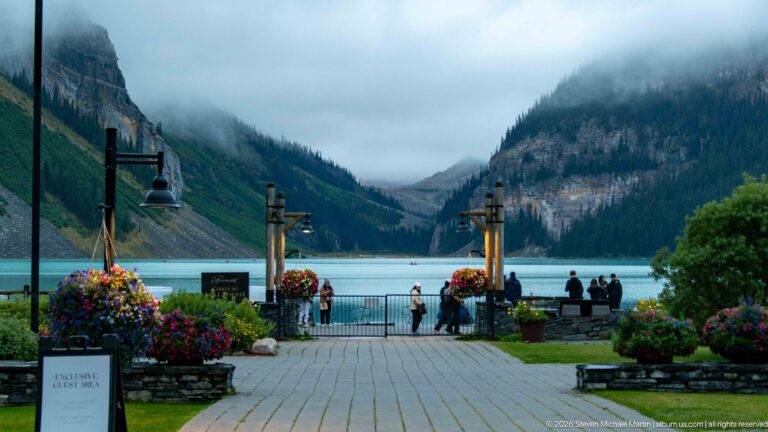 Visitors stand along Lake Louise’s paved promenade as flower baskets and lampposts lead toward the cloudy mountain-framed water.