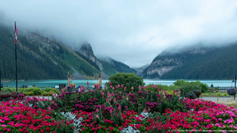 Pink and red flowers fill the foreground below turquoise Lake Louise and cloud-covered mountain slopes.