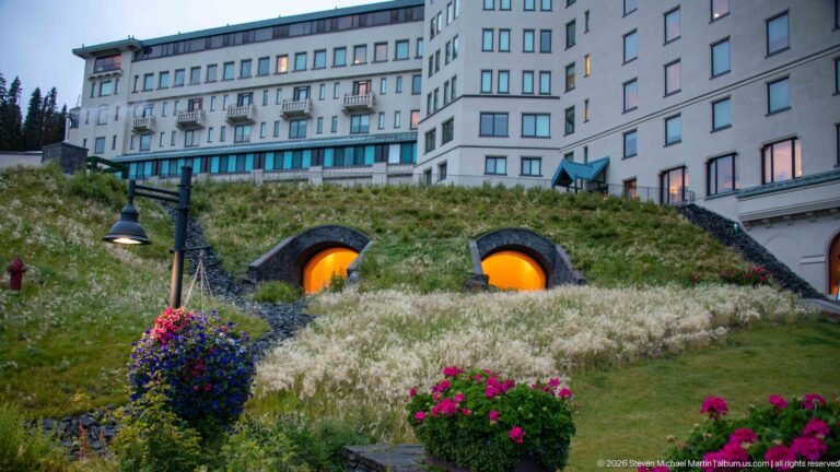 Two amber-lit arches sit in a flowered, grass-covered terrace below the pale façade of the Fairmont Chateau Lake Louise.