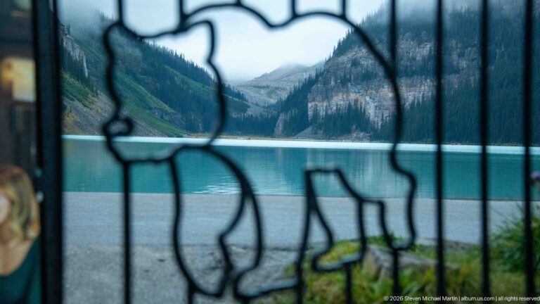 Out-of-focus iron scrollwork frames turquoise Lake Louise and forested mountain slopes beyond a lakeside walkway.
