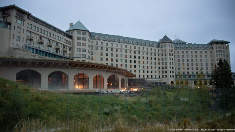 The Fairmont Chateau Lake Louise stands behind a rough garden slope, with warm lights glowing beneath its arched ground floor.